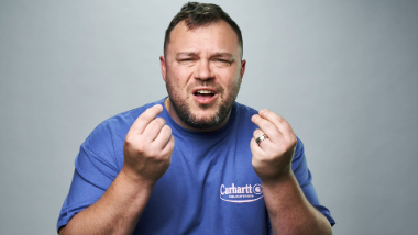Studio portrait of a person wearing a blue Carhartt shirt, holding both hands up near the face with fingers pinched together against a plain gray background.