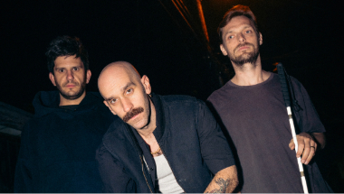 The X Ambassadors standing in a dark room. The one on the fair right is holding a stick. The one in the middle is bald. 